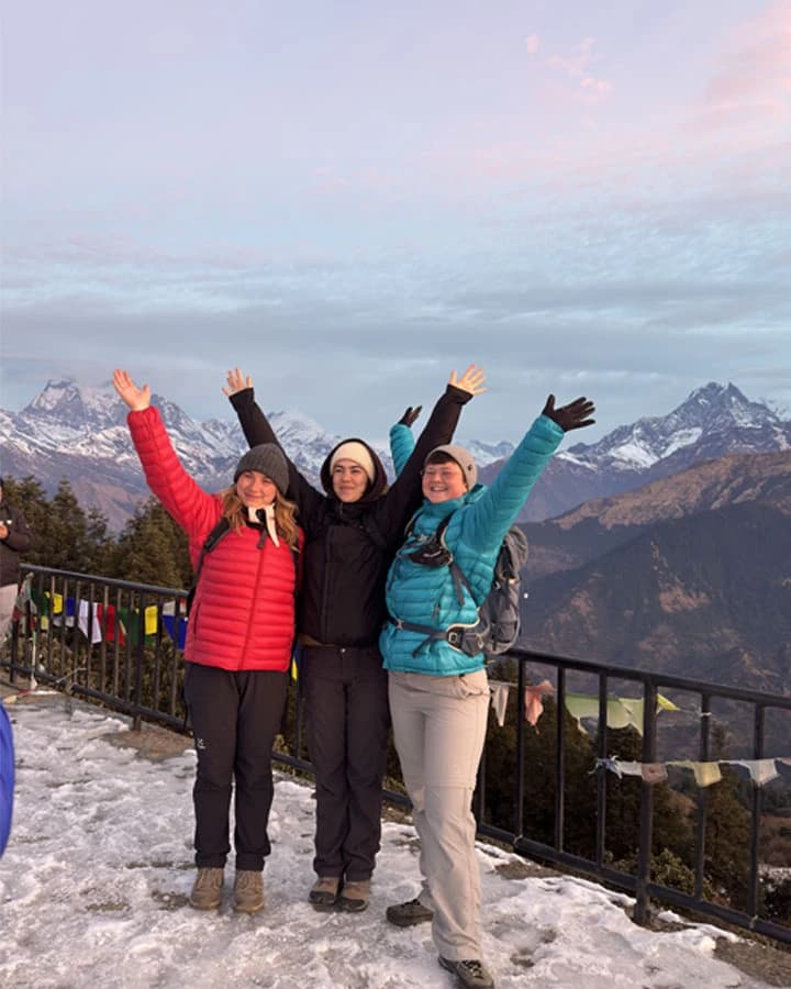 Three trekkers with raised arms celebrate at a snowy mountain viewpoint overlooking the Himalayan peaks.
